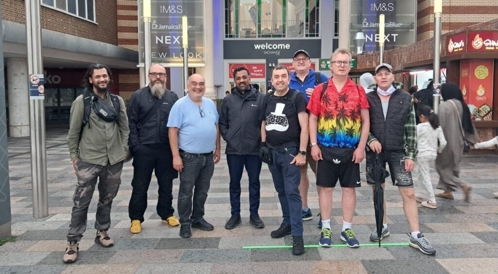 A group of men dressed in sports attire looking at the the camera and smiling outside the Ilford Station entrance of the Exchange Shopping Centre.