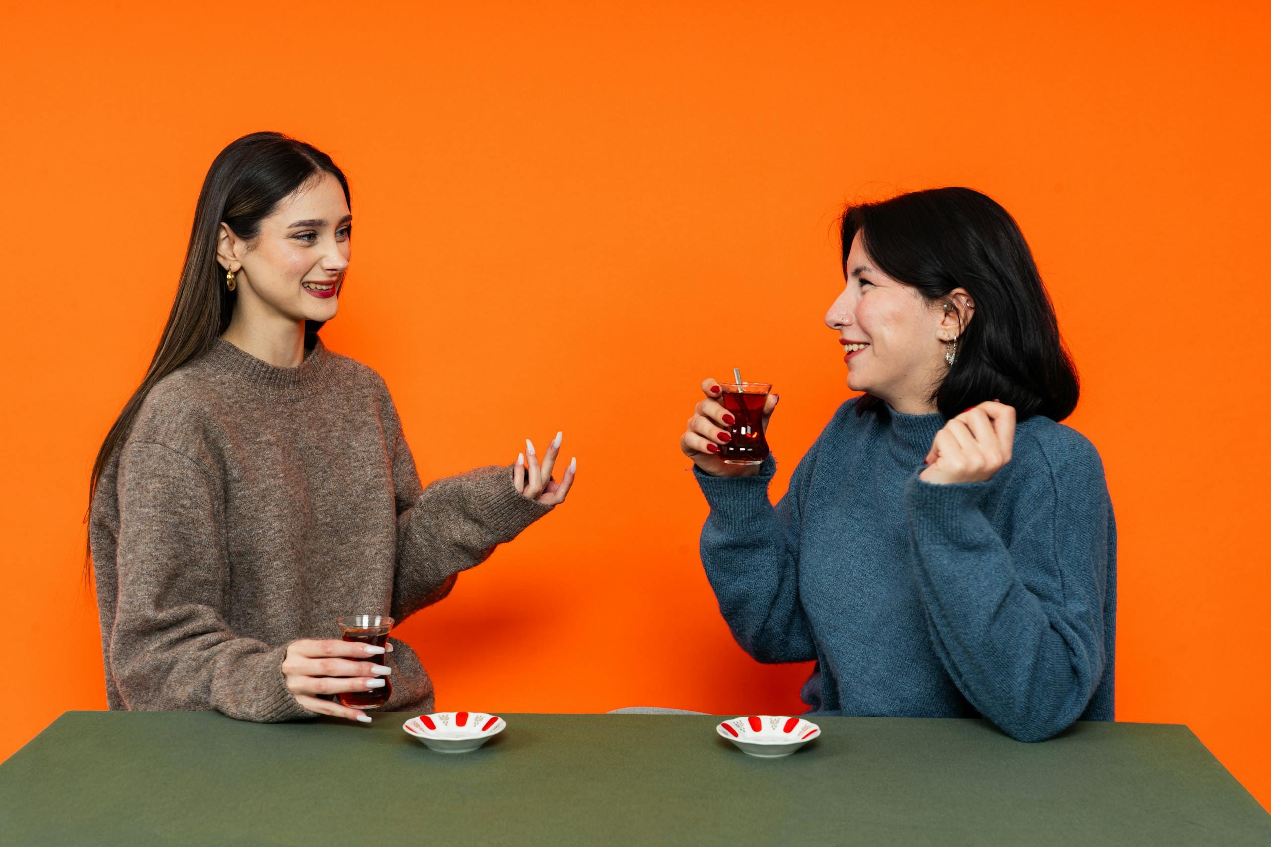 Two women enjoying Turkish tea and conversation indoors against a vibrant orange background.